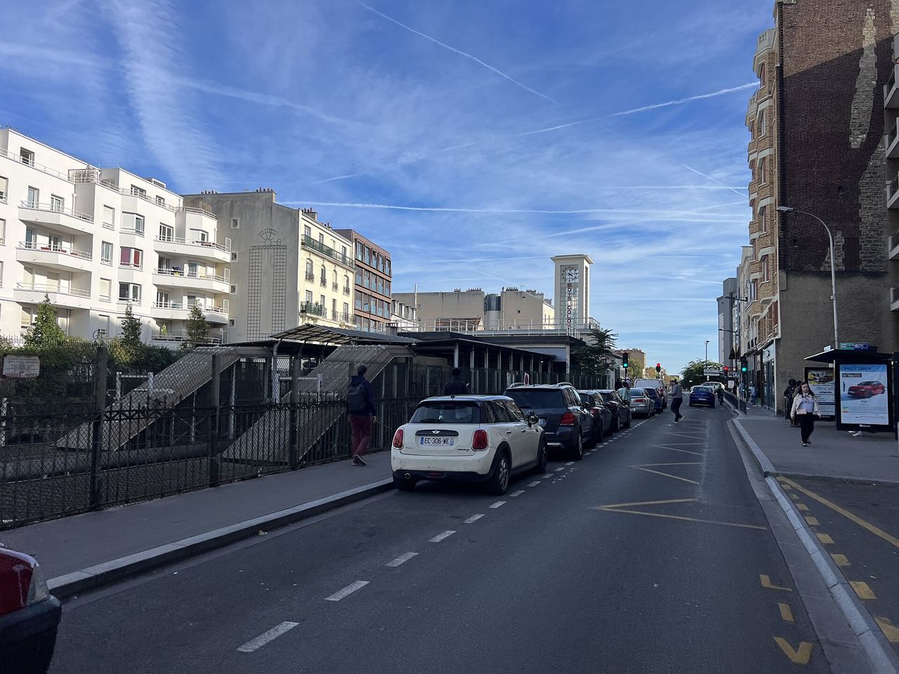 Rue longeant la gare de Bois-Colombes, avec un escalier d'accès aux quais, des voitures garées et un arrêt de bus