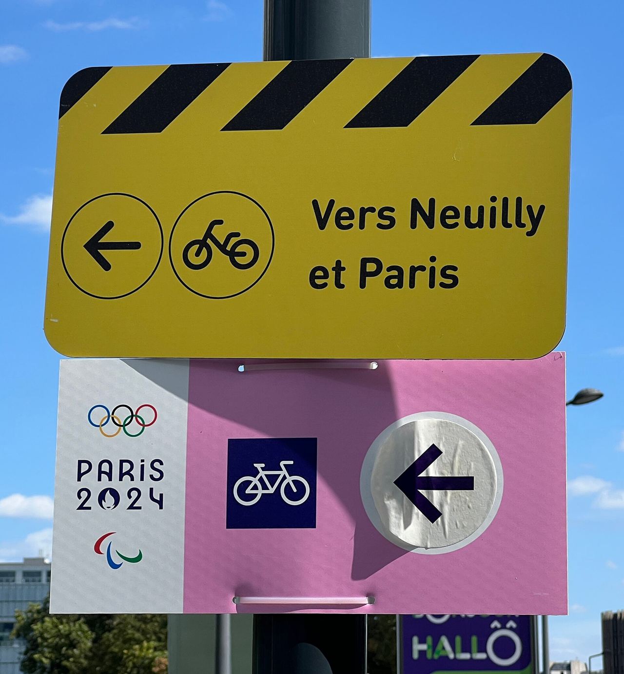 Panneau jaune de jalonnement cyclable « Vers Neuilly et Paris » au-dessus d'un panneau vélo des Jeux de Paris 2024, boulevard de Neuilly à Courbevoie.