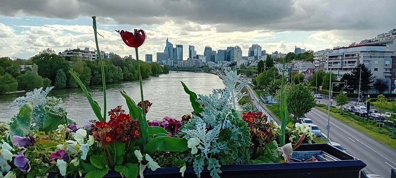 Jardinières fleuries sur le parapet du pont de Levallois, avec la Seine, les quais de Courbevoie et les tours de La Défense sous un ciel nuageux.