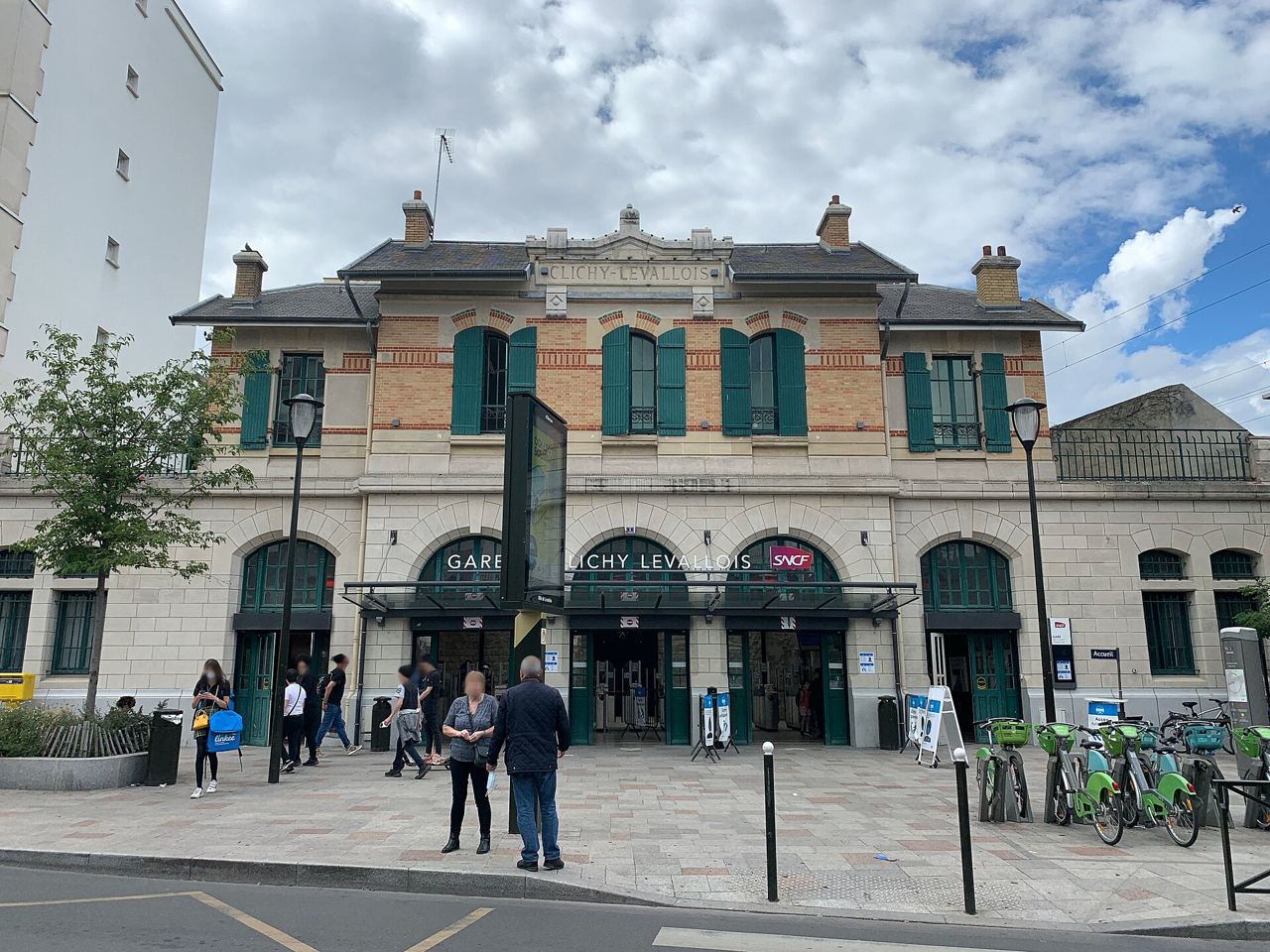 La façade de brique et de pierre de la gare de Clichy-Levallois, avec une station Vélib' et ses vélos verts devant l'entrée.