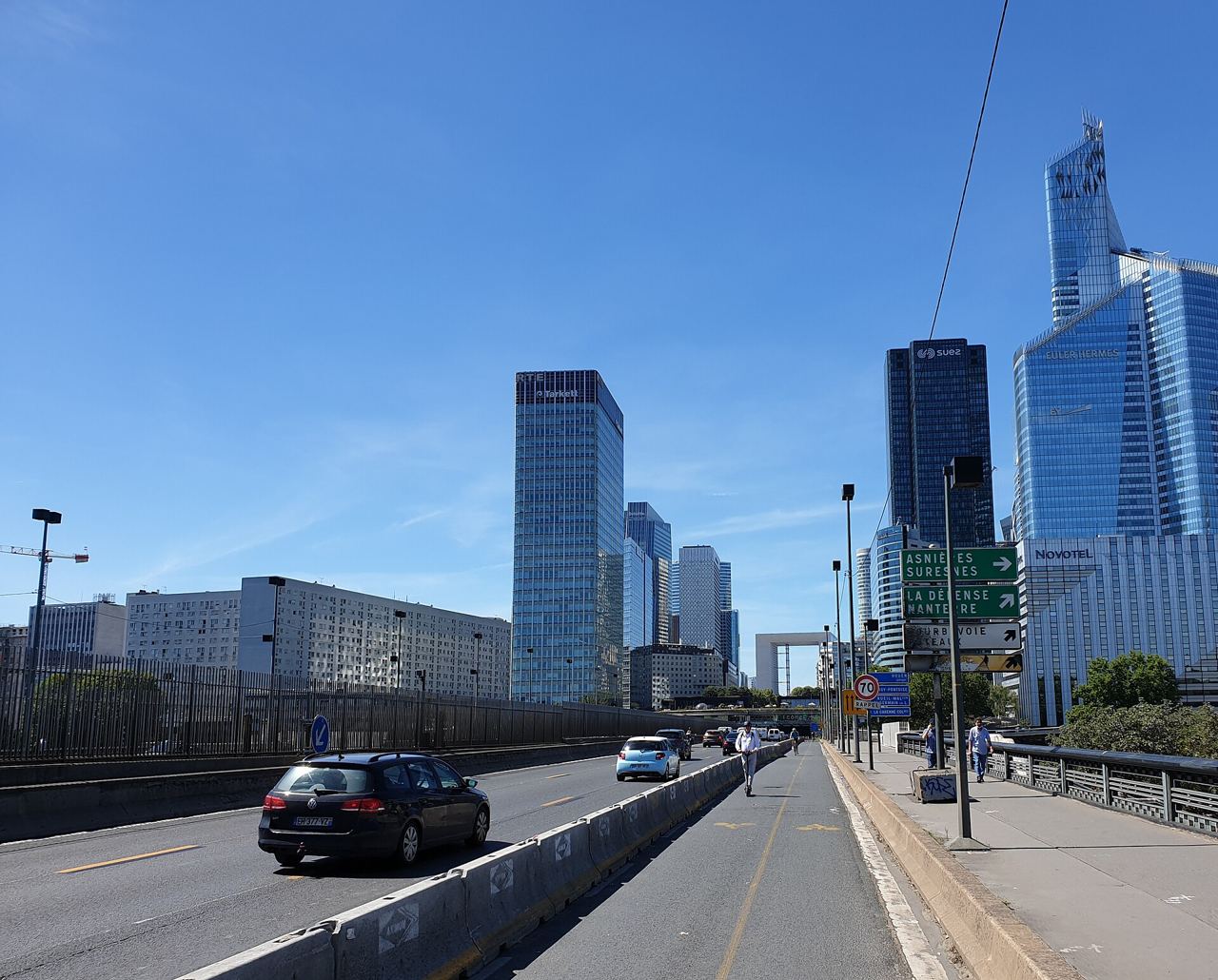 La piste cyclable provisoire du pont de Neuilly en 2020 : une voie de la RN13 séparée des voitures par des blocs de béton, vers les tours de La Défense, sous un ciel bleu.