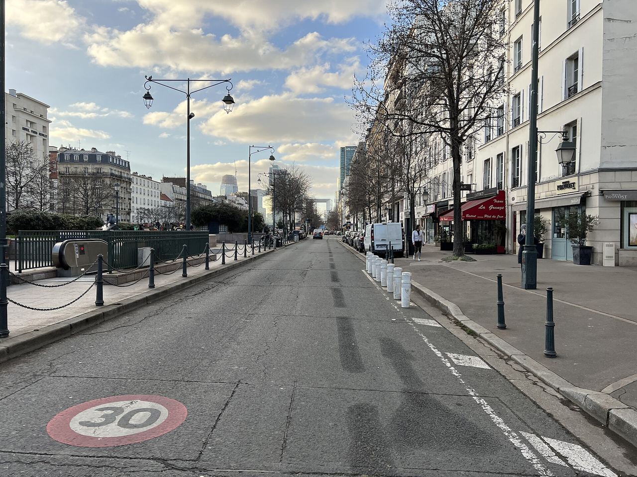 Contre-allée de l'avenue Charles-de-Gaulle à Neuilly-sur-Seine : un marquage 30 peint sur la chaussée, des potelets le long du trottoir et des immeubles en pierre sous un ciel nuageux.