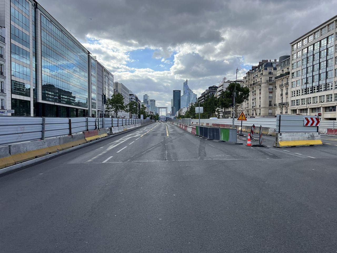 L'avenue Charles-de-Gaulle à Neuilly-sur-Seine pendant le chantier des Allées de Neuilly, en 2023 : chaussée vide bordée de barrières de travaux, et les tours de La Défense au bout de la perspective.
