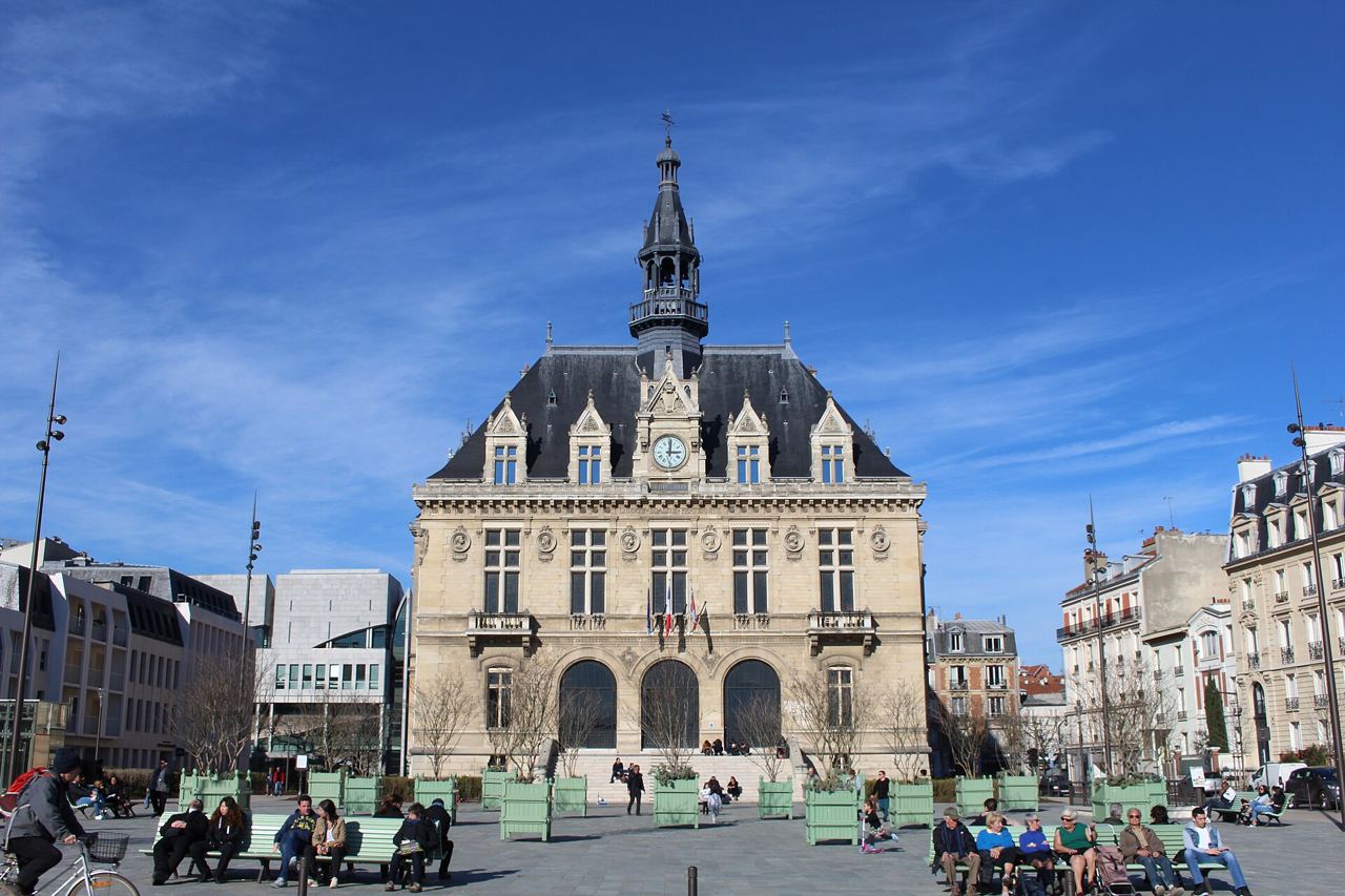 L'hôtel de ville de Vincennes sous un ciel bleu, avec son campanile et des passants assis sur la place devant la façade.
