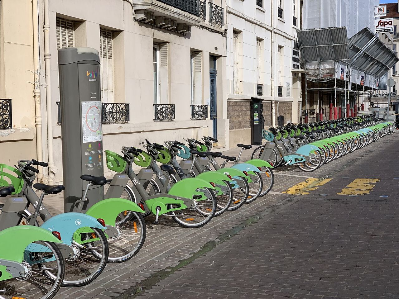 La station Vélib' de la gare RER de Vincennes, une longue rangée de vélos verts et bleus garés le long d'un trottoir pavé.